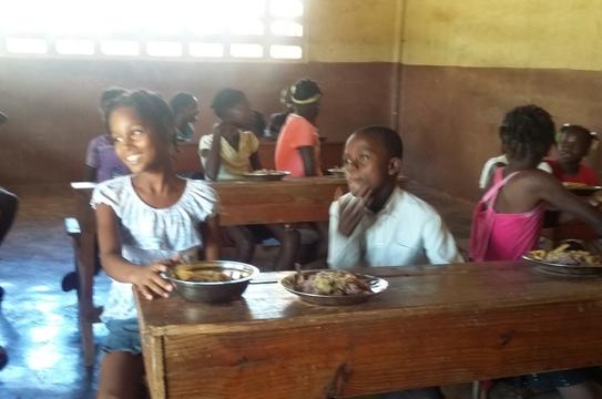 Haitian school children sitting at wooden desks eating a sponsored lunch